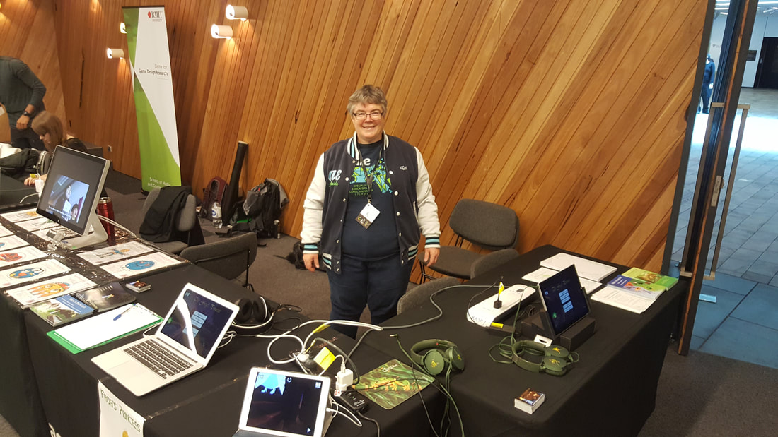 Kathy stands behind 2 tables with a Mac computer, a PC computer, an iPad and an Android phone with Frog's Princess playing on all of them.