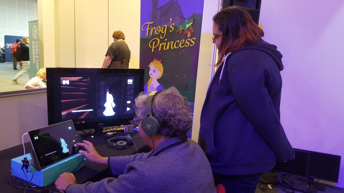 One person plays Frog's Princess on a small computer while the game play is reflected on a larger screen beside a banner saying 