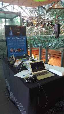 Kathy stands behind her display featuring a Mac computer, a PC computer, an iPad and an Android phone with Frog's Princess playing on all of them.