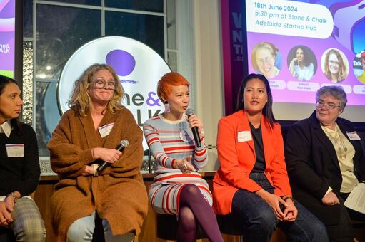 5 women sit on high stools in front of a Stone & Chalk venue logo with a powerpoint slide to the right showing their faces and names.  The woman on the left looks thoughtful, the next woman is holding a microphone looking humorous, the next woman, also holding a microphone, is speaking with great charm, the next woman is looking up as she listens, the woman on the right is looking across to watch the speaker.