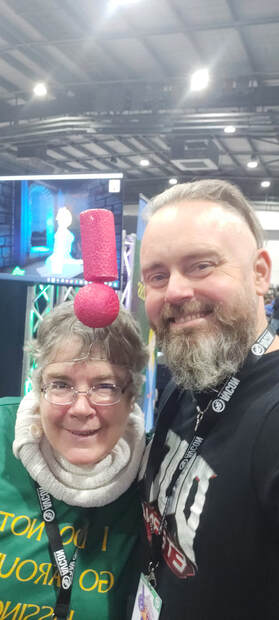 Kathy wears a red foam exclamation mark as a hat and an AVCon lanyard.  Her shirt has mirror writing incompletely saying I do not go around kissing frogs.  Beside her a bearded man smiles and holds up the camera.