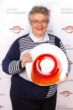 Kathy holds artistically painted plate in front of a display board decorated with Women In Innovation logos